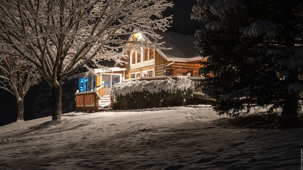 A snow-covered landscape with a wooden house featuring a visible porch and a well-lit entrance.