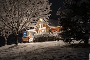 A snow-covered landscape with a wooden house featuring a visible porch and a well-lit entrance. - Western Catskills Paradise - Four-Season Mountaintop Retreat (Bainbridge)