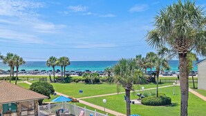Ein Poolbereich mit Liegestühlen und Sonnenschirmen, eine Rasenfläche mit Palmen und Blick auf das Meer und geparkte Autos.