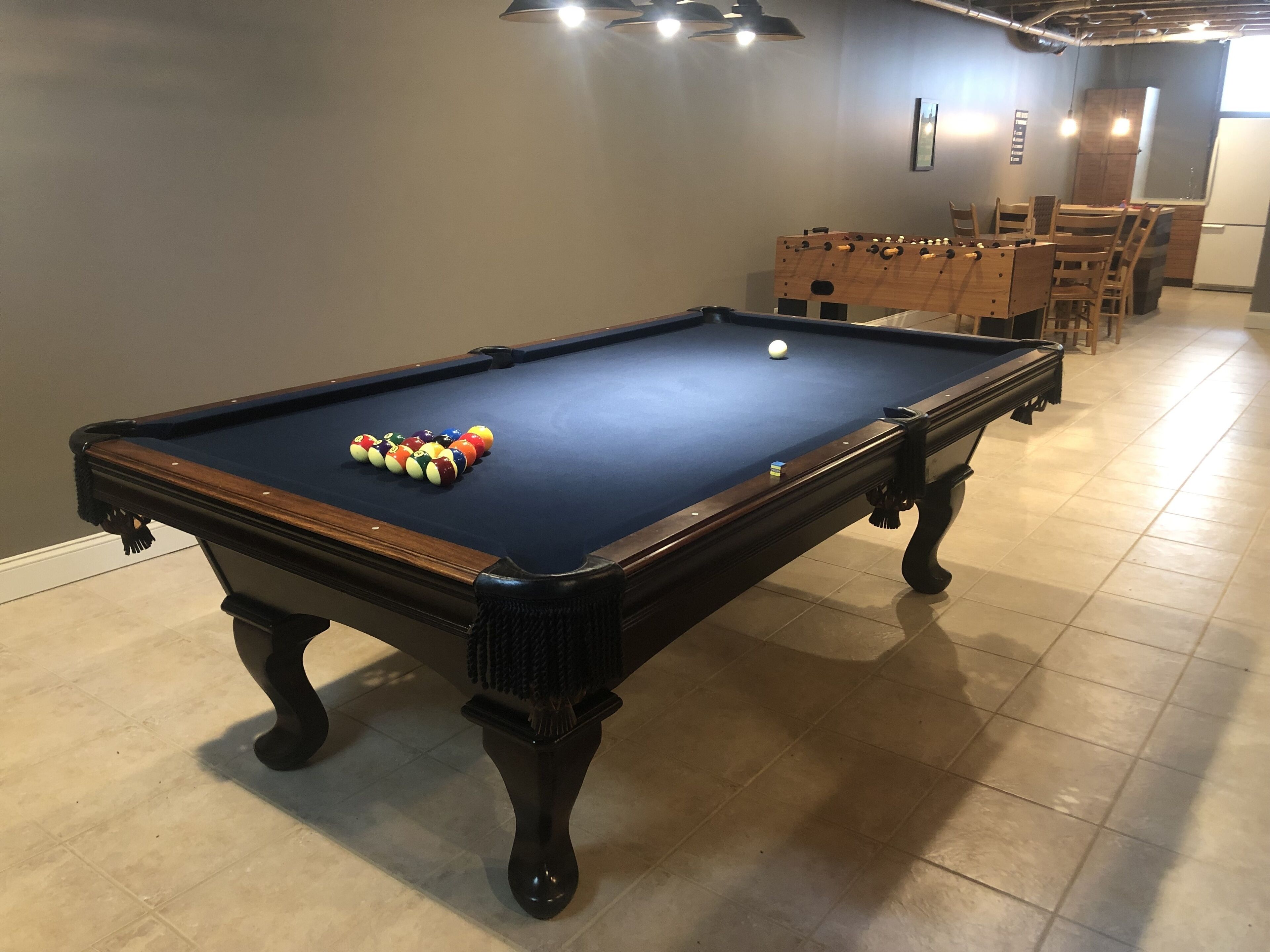 A pool table with balls and a wooden table in a room with tiled flooring and a dining area in the background.