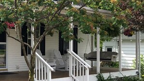 Een wit huis met een veranda, twee Adirondack-stoelen, een barbecue en een kleine boom met gele bloemen.