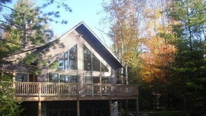 A two-story wooden cabin with a balcony, surrounded by trees and a stone wall.