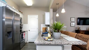 A modern kitchen with a stainless steel refrigerator, oven, and microwave. There is a granite countertop with a potted plant and a bottle of wine. A wicker sofa is visible in the background.