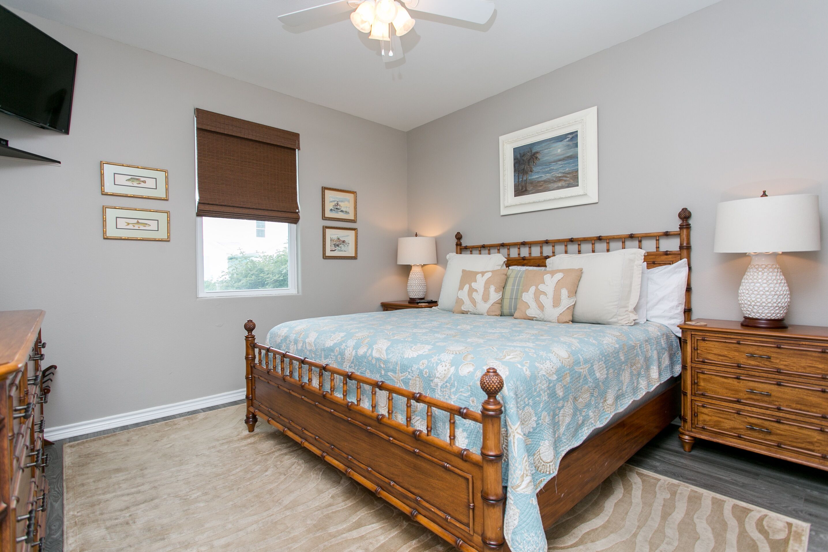 A bedroom with a wooden bed, two bedside tables with lamps, a ceiling fan, a window with blinds, and framed pictures on the walls.