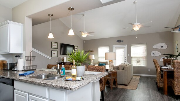 A modern kitchen with granite countertops, white cabinetry, and a central island. There are ceiling fans, pendant lights, and a dining area with wicker furniture.