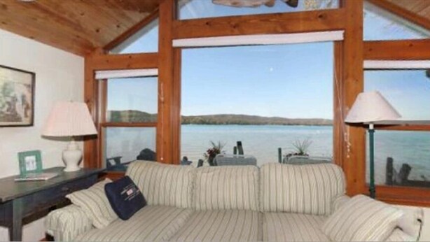A room with a large window, a sofa, a lamp, and a framed picture on the wall. - Glen Lake  - Spectacular Dune Views & Complete Privacy! (Glen Arbor)