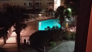 Vue nocturne d’un hôtel avec une piscine, des palmiers et un immeuble à plusieurs étages en arrière-plan.