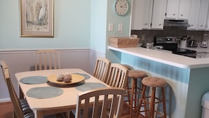 Un coin repas avec une table et des chaises en bois, un ventilateur de plafond, et une cuisine dotée de meubles et d’appareils électroménagers blancs.