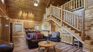 Interior de una cabaña de troncos con una escalera de madera, una sala de estar con sofá y mesa de centro, y una zona de cocina.