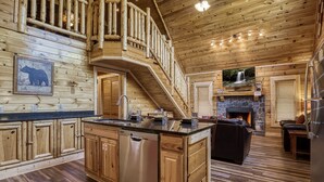 Interior de una cabaña con detalles de madera, una cocina con lavavajillas, chimenea y escalera que lleva a una zona superior.