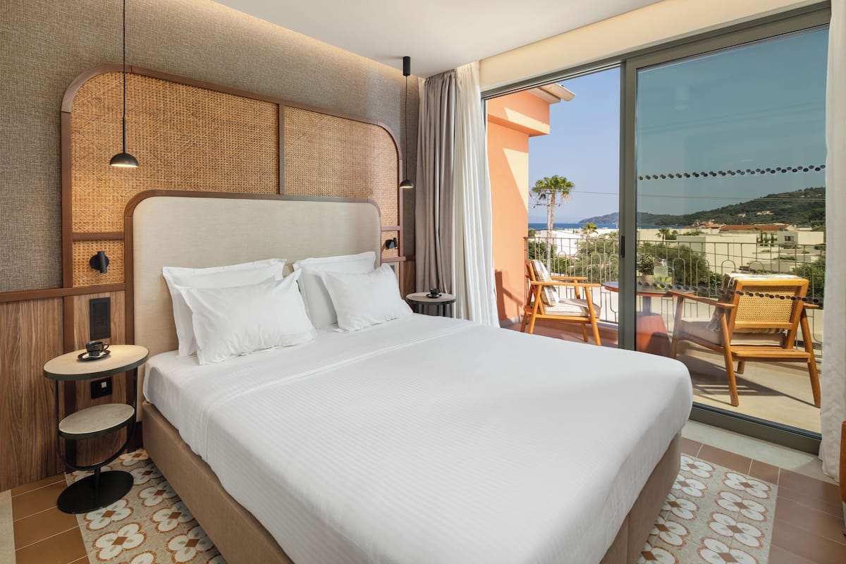 A hotel room with a large bed, a small table, and a balcony with a view of the sea.