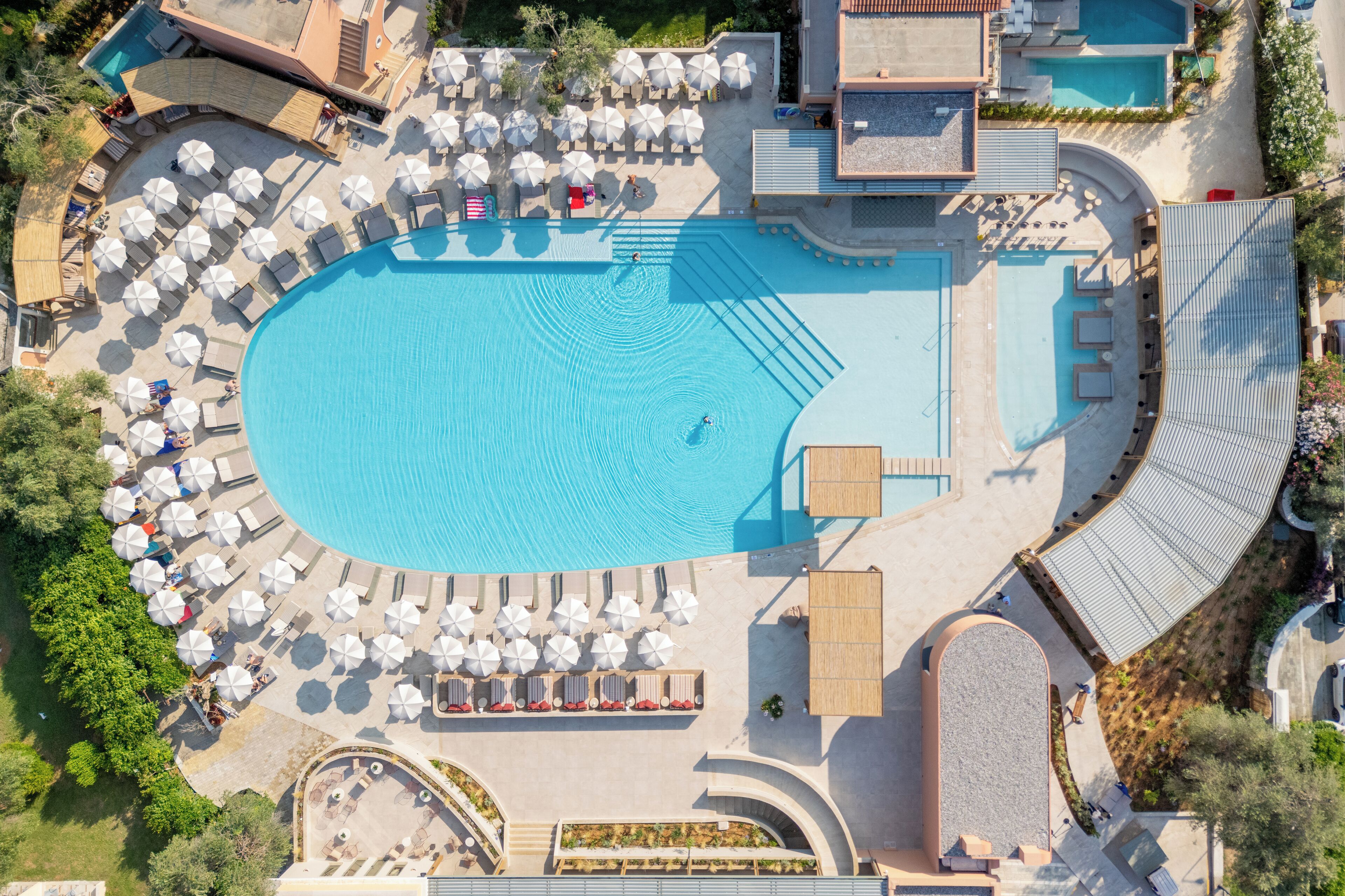 An aerial view of a resort with a large central swimming pool, multiple sun loungers, and umbrellas around the pool area.