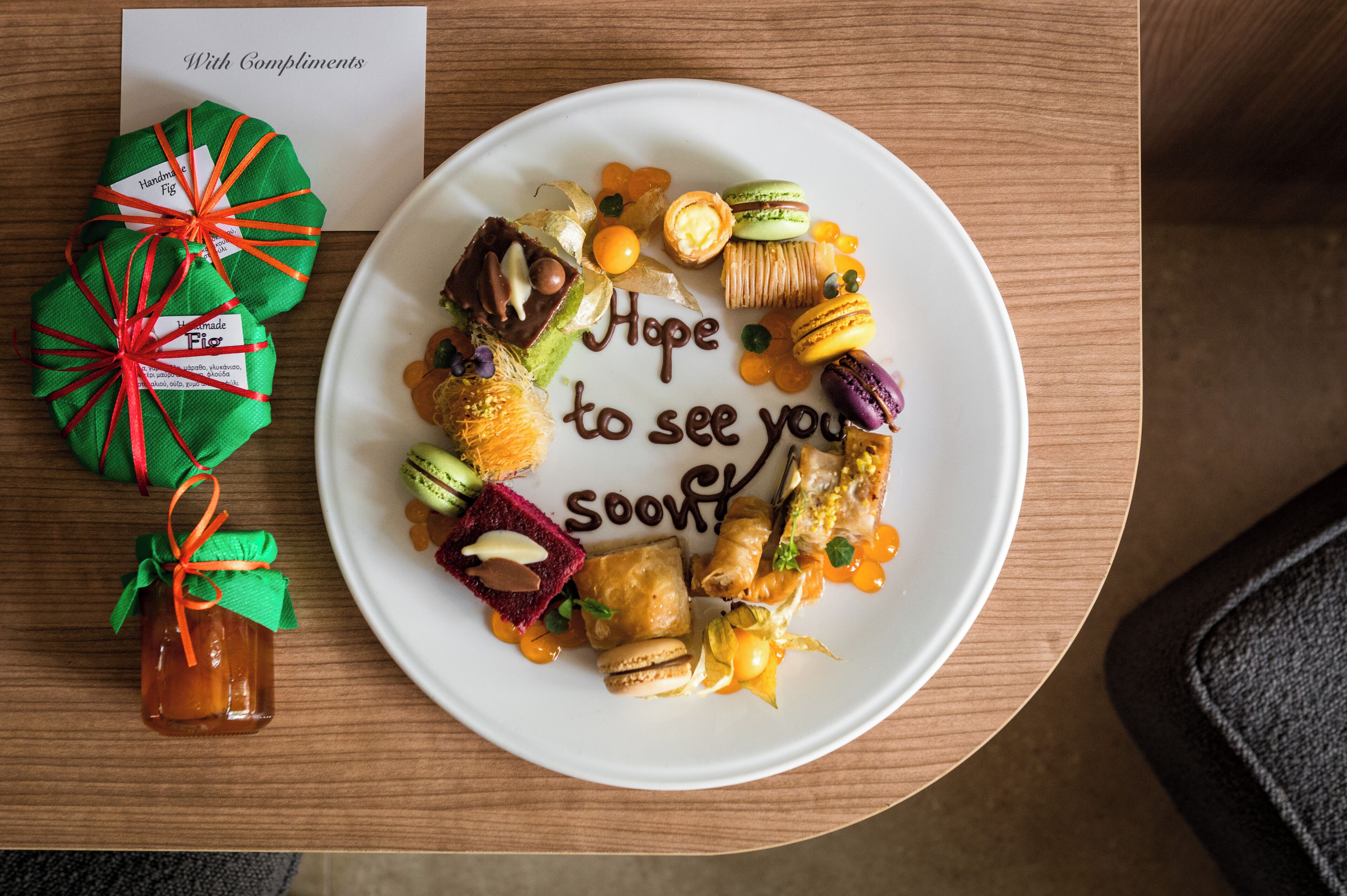 A plate of assorted pastries and chocolates with a message "Hope to see you soon" on a wooden table.