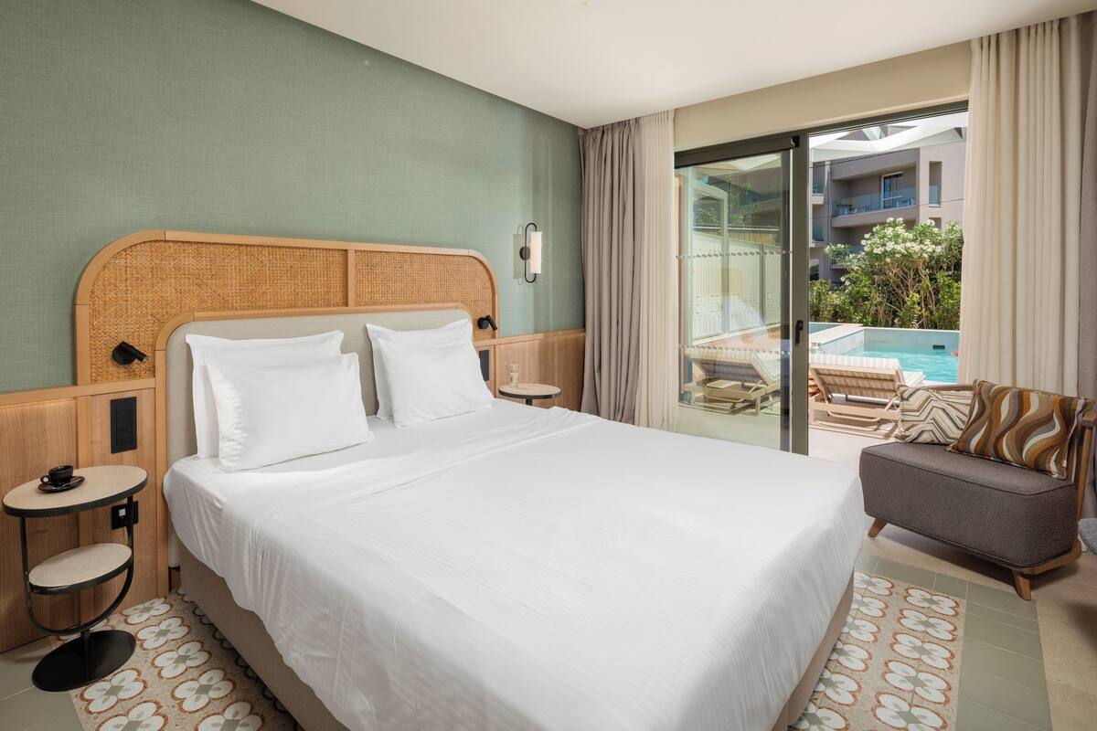 A hotel room with a large bed, a small round table, a sofa, and a view of a pool and building.