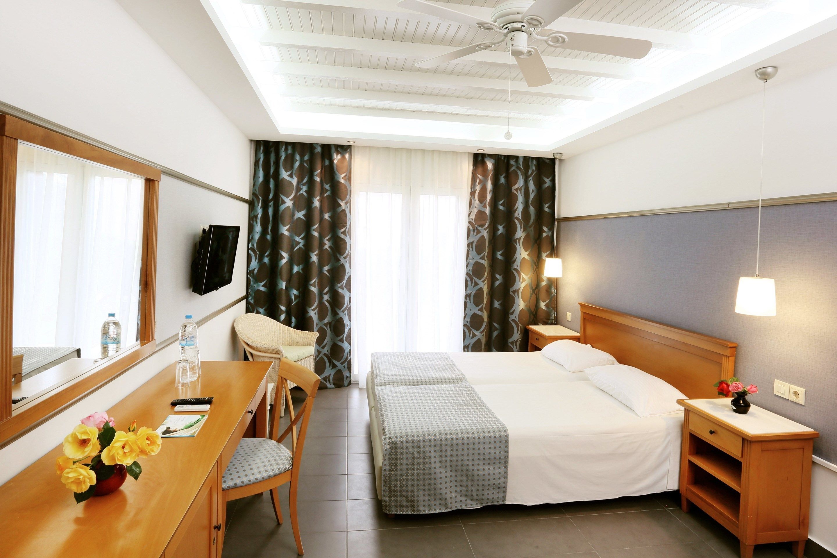 A hotel room with a bed, a desk with a chair, a bedside table, a ceiling fan, and curtains.