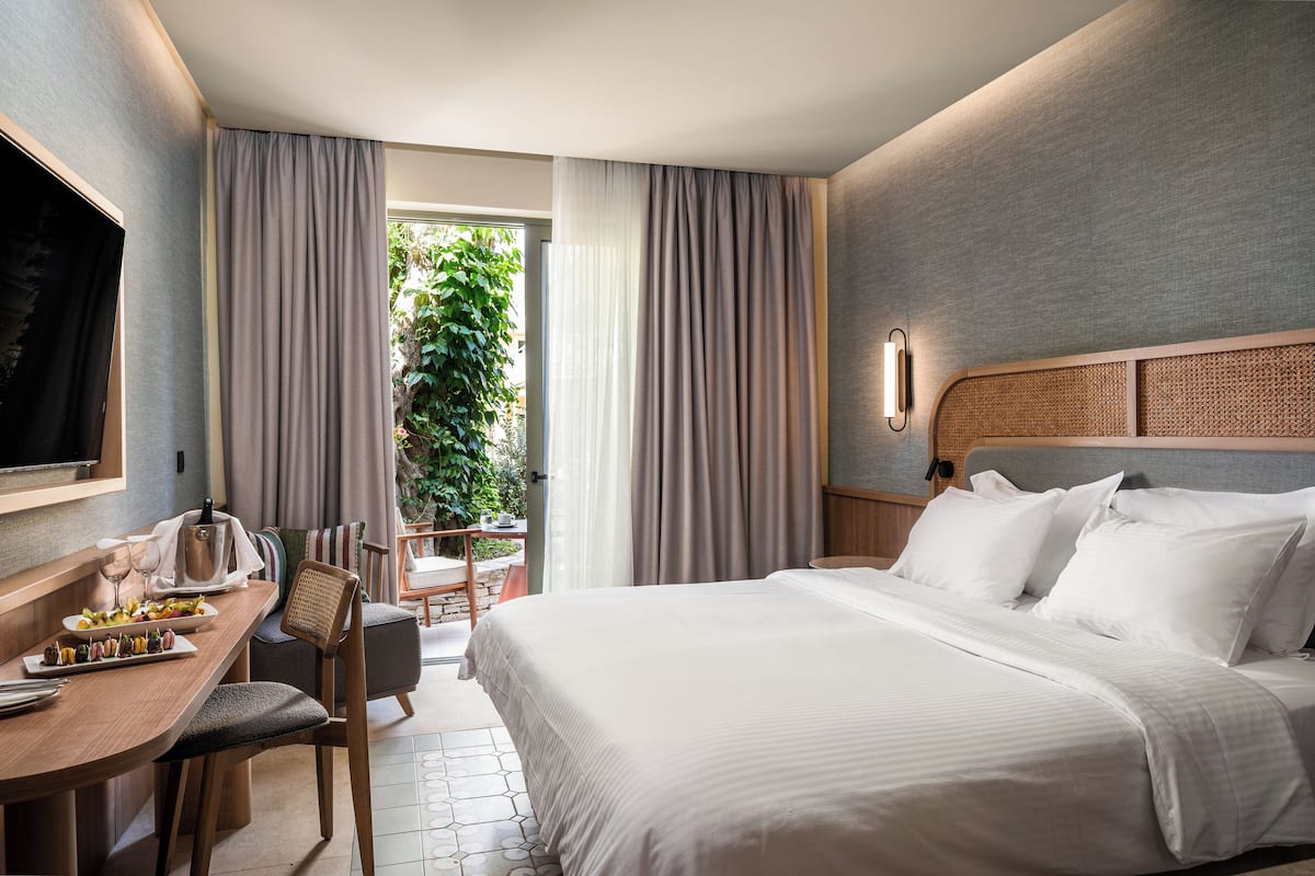 A hotel room with a bed, a desk with a chair, a television, and a view of greenery outside.