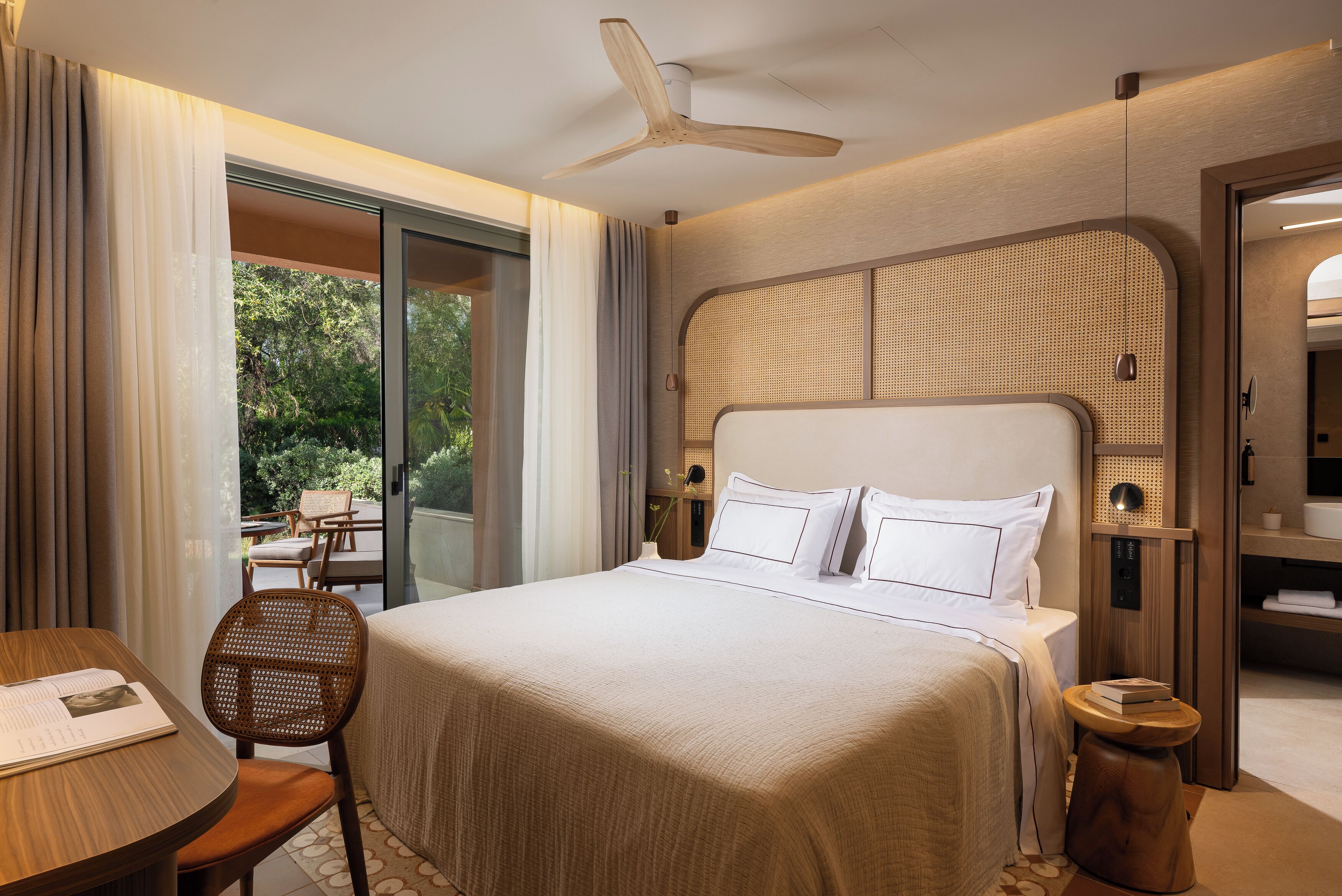 Habitación de hotel con una cama grande, ventilador de techo, zona de estar con una mesa y sillas, y vistas al exterior.