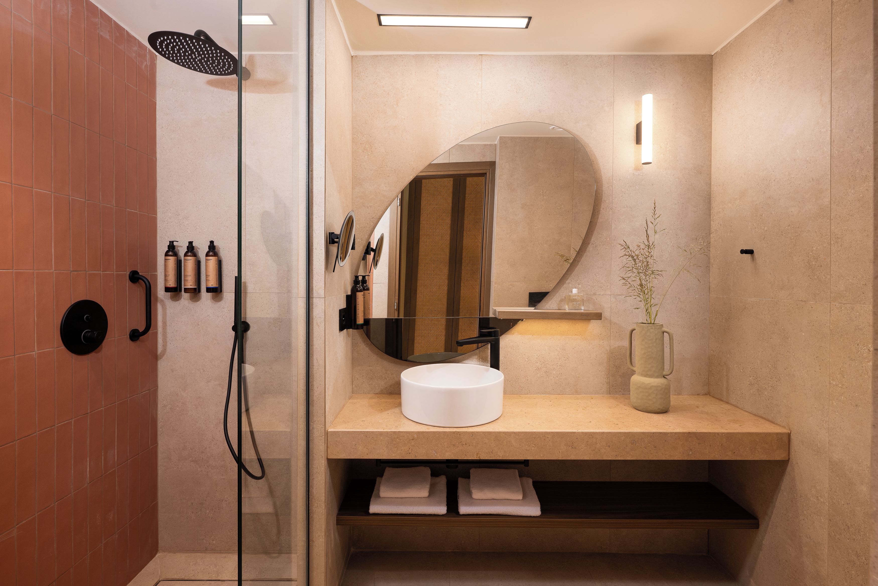 A modern bathroom with a glass-enclosed shower, a round mirror, a sink with a vase, and toiletries on the wall.