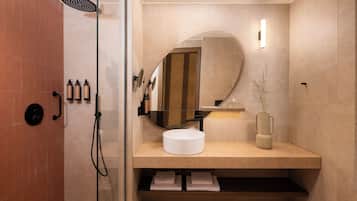 A modern bathroom with a glass-enclosed shower, a round mirror, a sink with a vase, and toiletries on the wall.