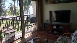 Sala de estar com uma televisão grande, sofá, poltrona e varanda com vista para palmeiras e praia.