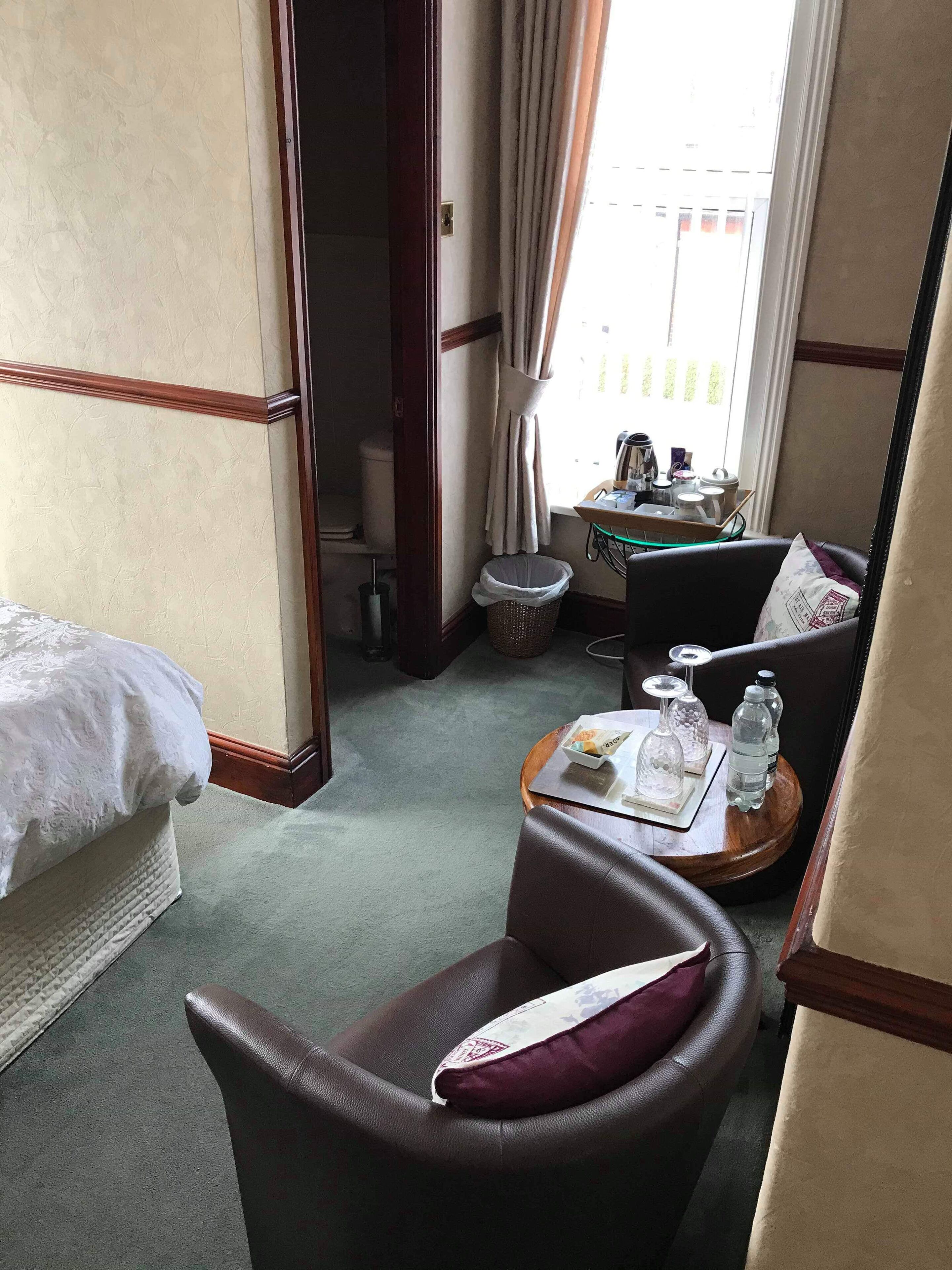 Une chambre d’hôtel avec un lit, une chaise, une petite table avec un plateau, une poubelle et une fenêtre avec des rideaux.