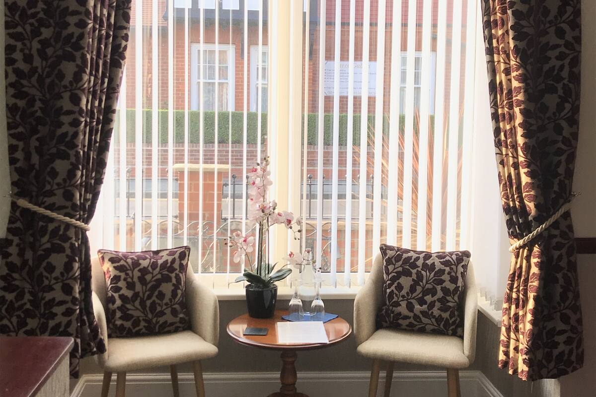 Ein Zimmer mit zwei Sesseln, einem kleinen Tisch und einem Fenster mit vertikalen Jalousien.