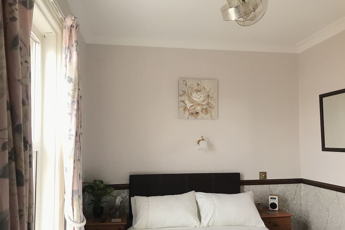 Ein ordentlich eingerichtetes Schlafzimmer mit einem Bett, Nachttischen, einer Wandlampe, einem gerahmten Blumenbild und einem Fenster mit Vorhängen.