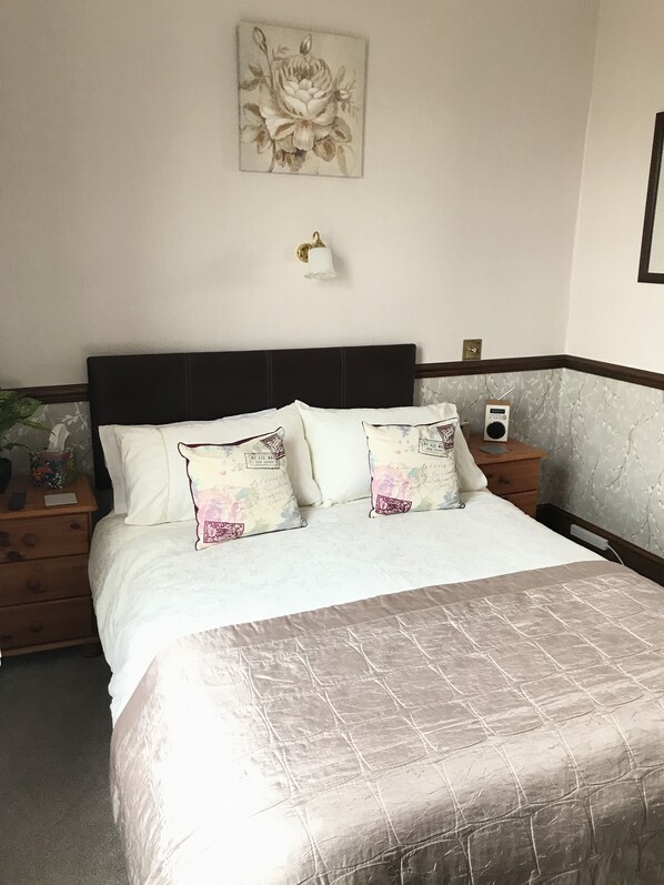 A neatly made bed with a quilted headboard, two bedside tables with lamps, a radio, and a framed artwork on the wall.