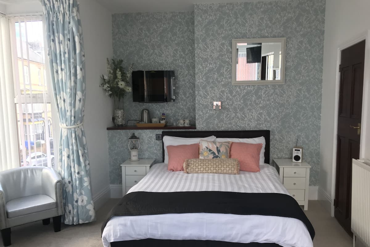 Ein Schlafzimmer mit Bett, Nachttischen, einem Sessel, einem Spiegel und einem Fenster mit Vorhängen.