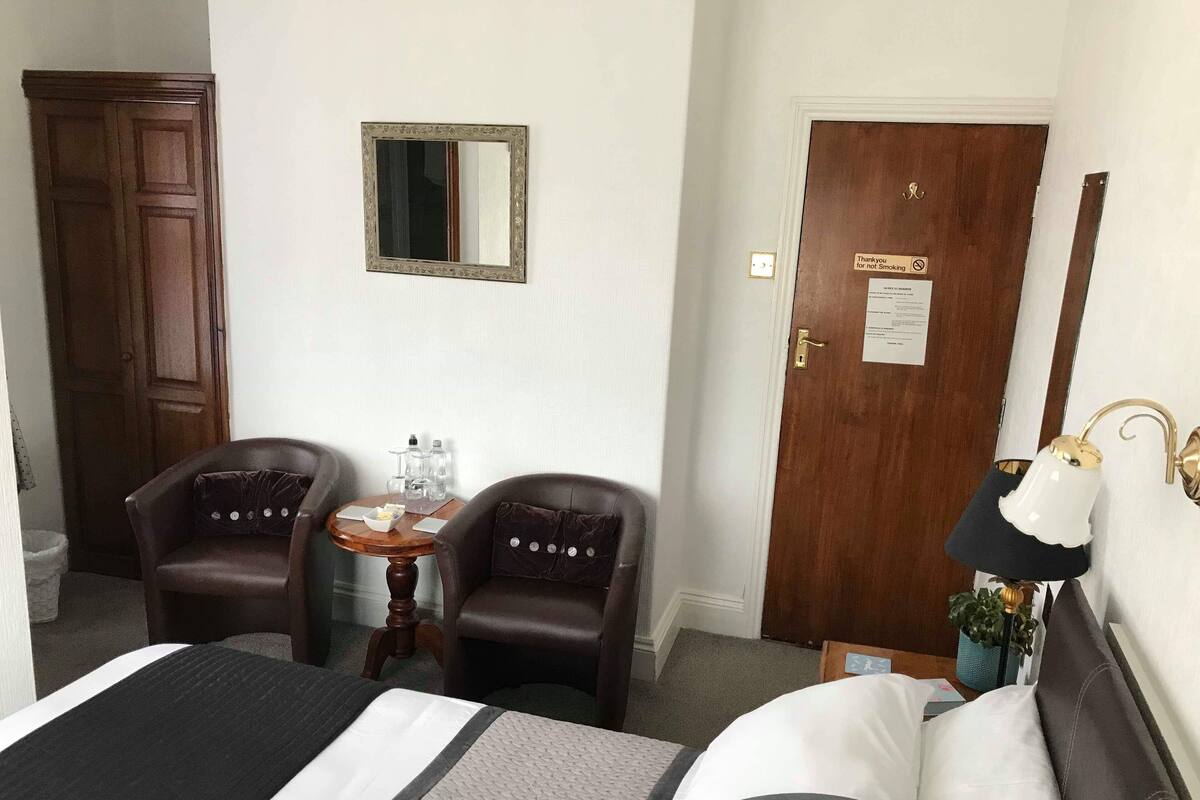 Ein Hotelzimmer mit einem Bett, zwei Sesseln, einem kleinen Tisch mit einer Wasserflasche, einem Spiegel und einer Tür mit einem Rauchverbot-Schild.