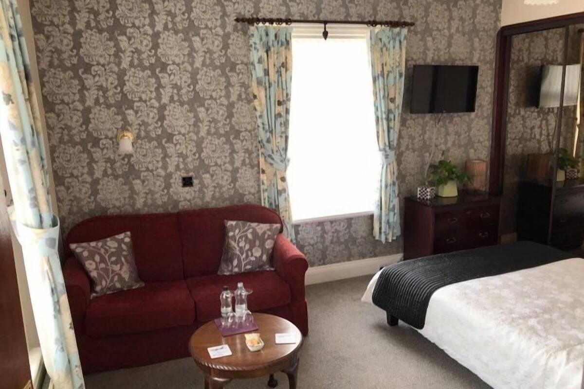 Ein Hotelzimmer mit einem roten Sofa, einem runden Holztisch, einem Bett mit gestreifter Bettdecke und einem an der Wand befestigten Fernseher.