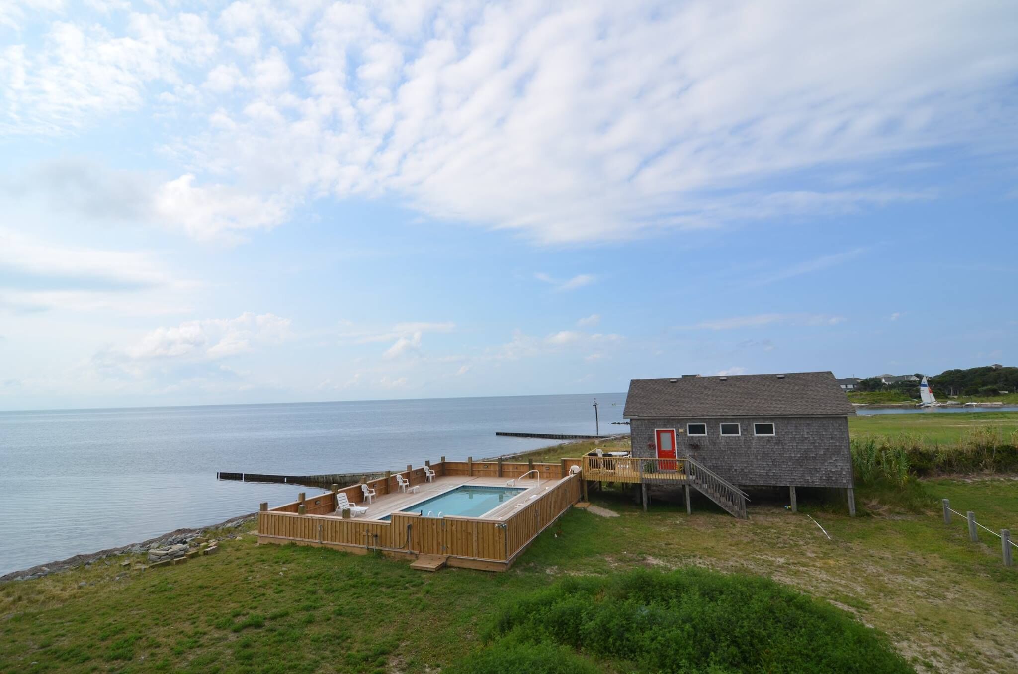 A wooden deck with a pool, a small house with a red door, and a view of the sea.