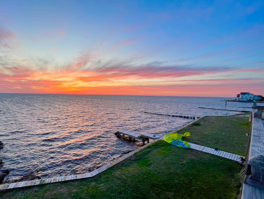 A coastal view with a wooden pier extending into the water, a grassy area, and a building in the background.