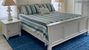 Un dormitorio con una cama blanca, ropa de cama a rayas, dos mesitas de noche con lámparas, una alfombra azul y un cuadro en la pared.