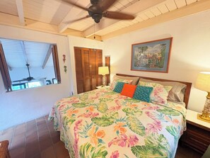 A bedroom with a bed, a ceiling fan, a window with blinds, a painting on the wall, and a nightstand with a lamp.