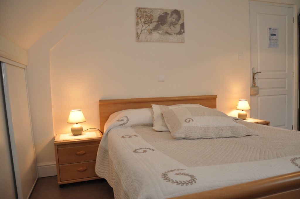 A neatly made bed with a white bedspread and pillows, two bedside lamps, a small wooden nightstand with a drawer, and a framed picture on the wall.
