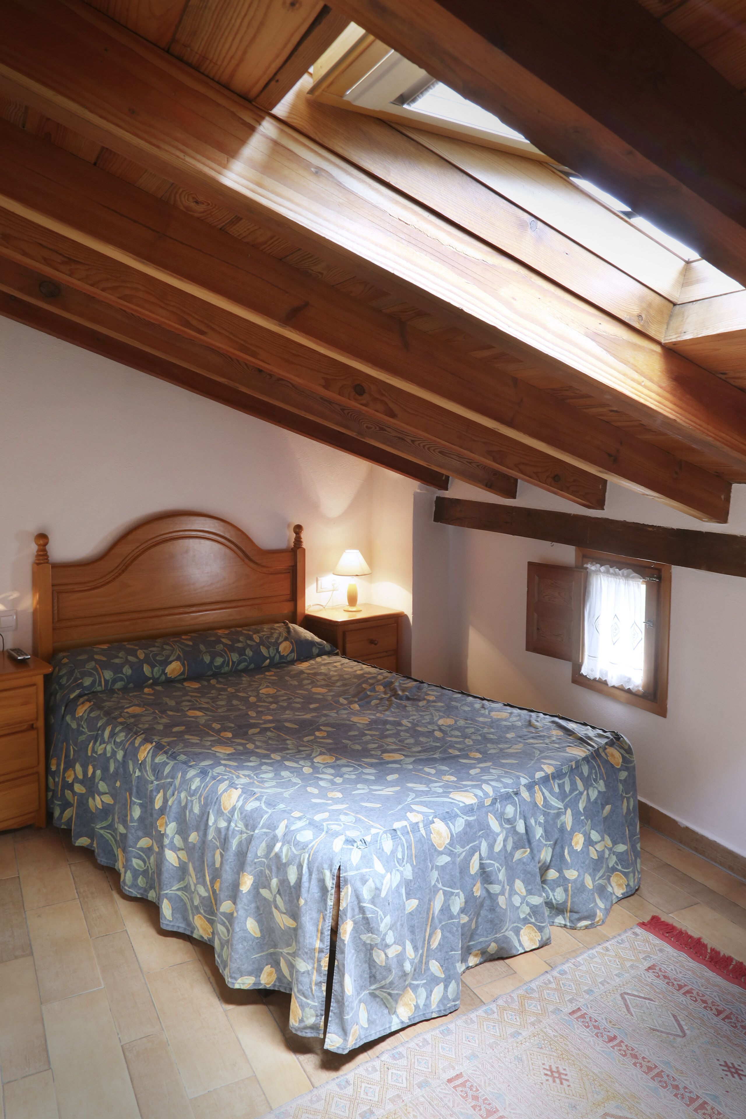 A bedroom with a wooden ceiling, a bed with a floral bedspread, a nightstand with a lamp, and a window with curtains.