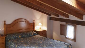 A bedroom with a wooden ceiling, a bed with a floral bedspread, a nightstand with a lamp, and a window with curtains.