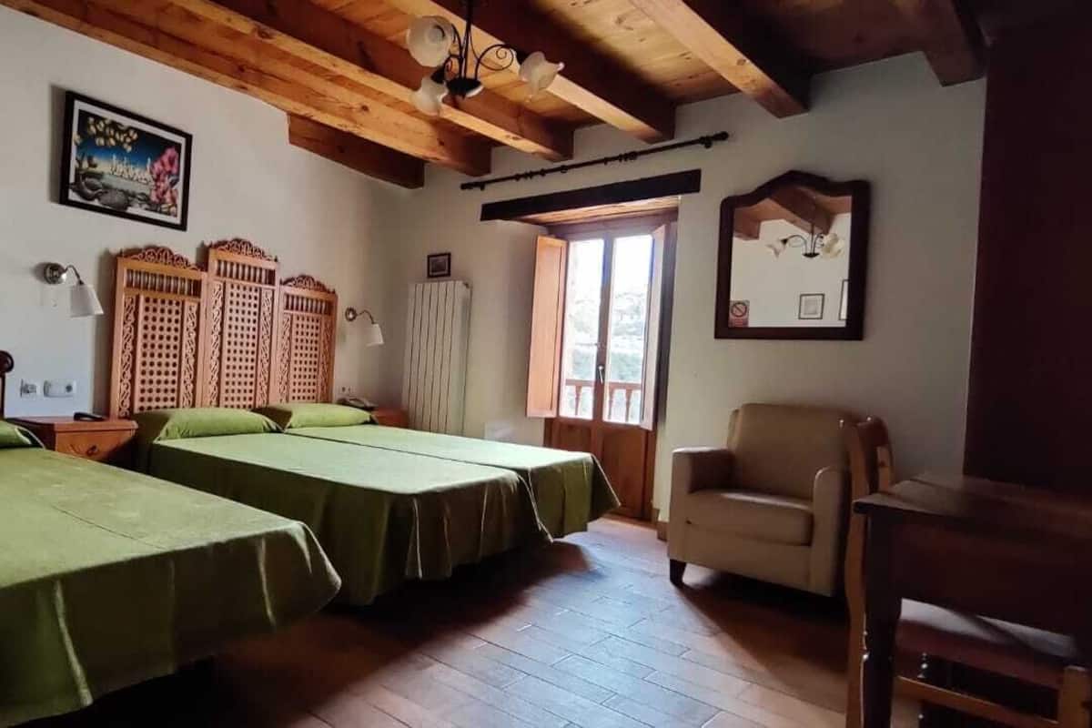 A room with two beds, wooden headboards, a wooden ceiling, a chair, and a window with a view.