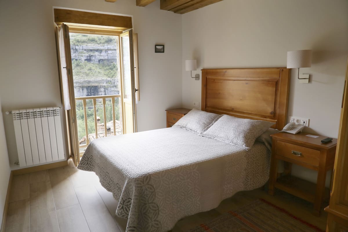 A bedroom with a wooden headboard, a bed with a patterned bedspread, two bedside tables with lamps, a radiator, and a view of a natural landscape through an open door.