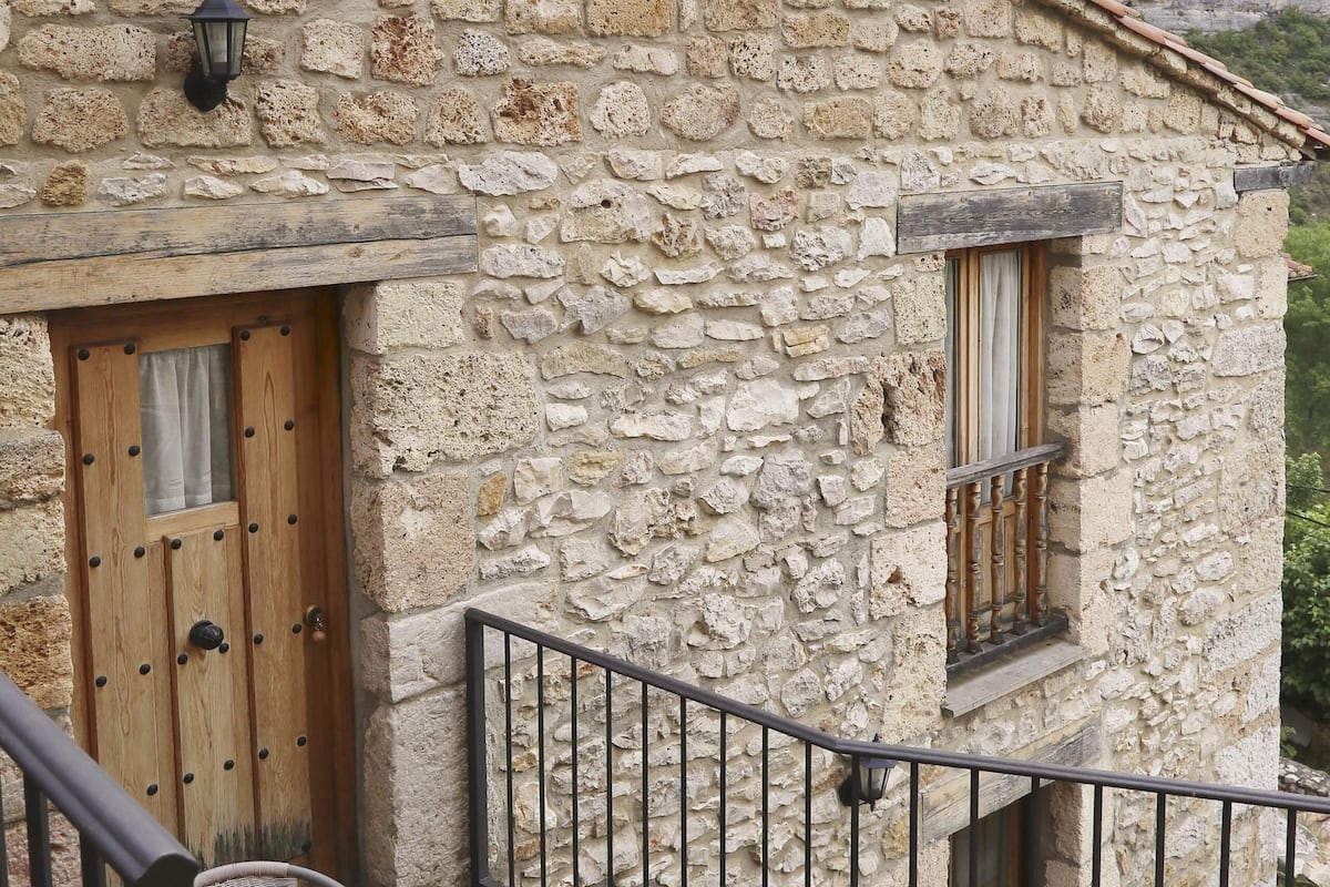A stone building with a wooden door and window, a balcony with metal railings, and wicker chairs.