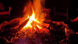 Un feu de camp avec de la fumée qui s’élève, une chaise et la silhouette d’un bâtiment se détachant sur un ciel nuageux.