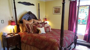 A four-poster bed with a red and gold patterned bedspread, flanked by two nightstands with lamps, a wooden headboard, and a window with red curtains. - Garfield Guest House - Hawksbill Suite (Luray)