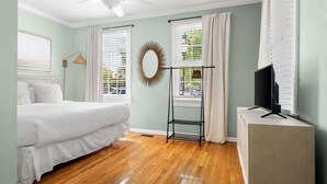A bedroom with a bed, a TV on a stand, a ceiling fan, and a window with curtains.