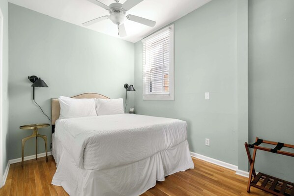 A bedroom with a bed, two bedside lamps, a ceiling fan, a window with blinds, and a wooden bench.