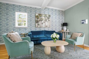 A living room with a blue patterned wallpaper, a blue sofa, two matching armchairs, a coffee table, and a painting of palm trees.