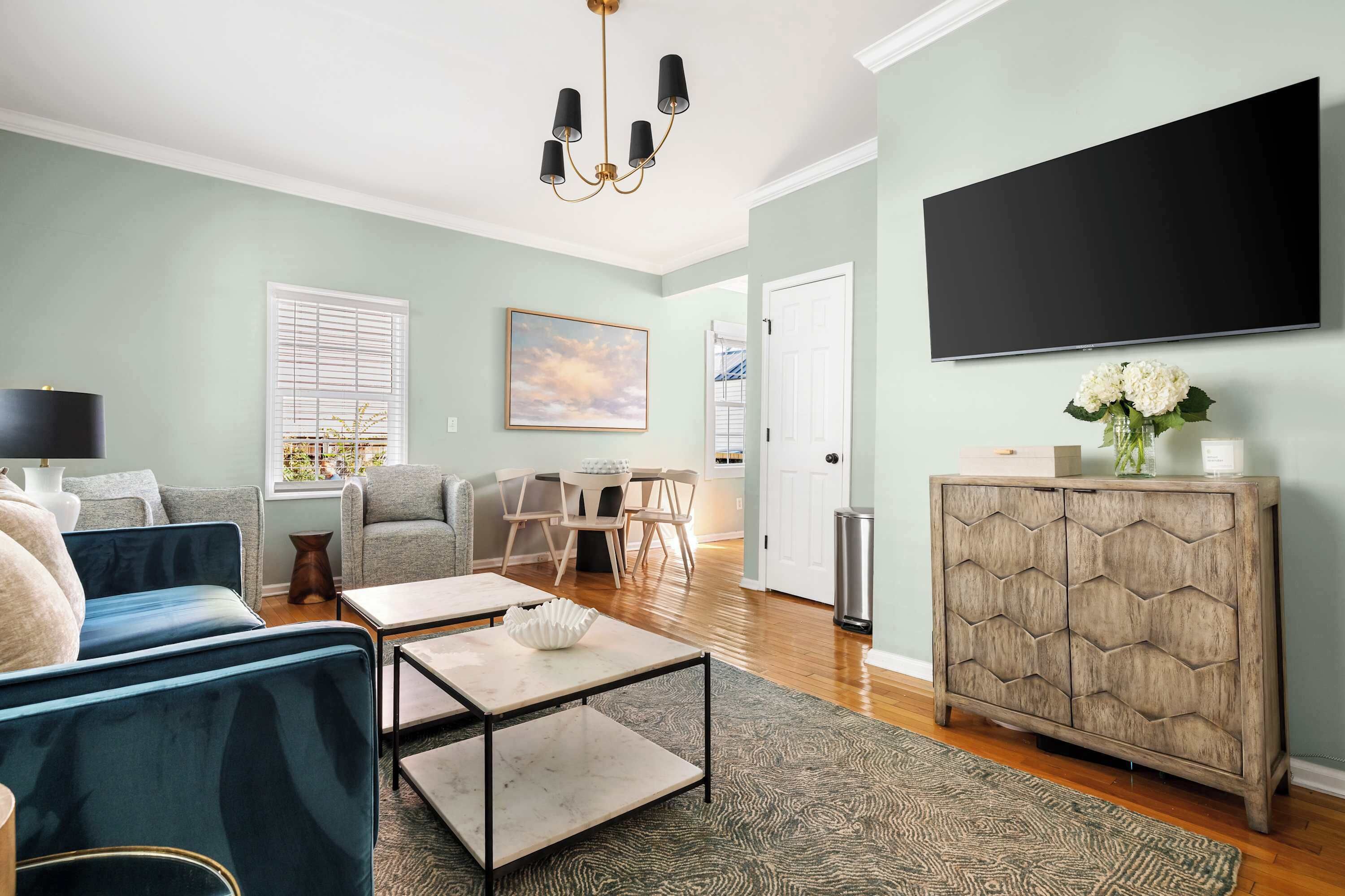 A living room with a sofa, coffee table, and a cabinet. There's a television mounted on the wall and a window with blinds.