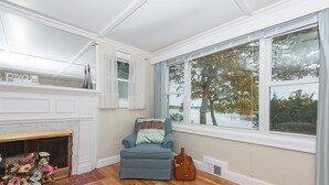 A room with a fireplace, a blue armchair, a large window with a view of trees, and a wooden floor. - Oceanfront Home Great For Families! Near Amenities, Canal, & More! (Buzzards Bay)