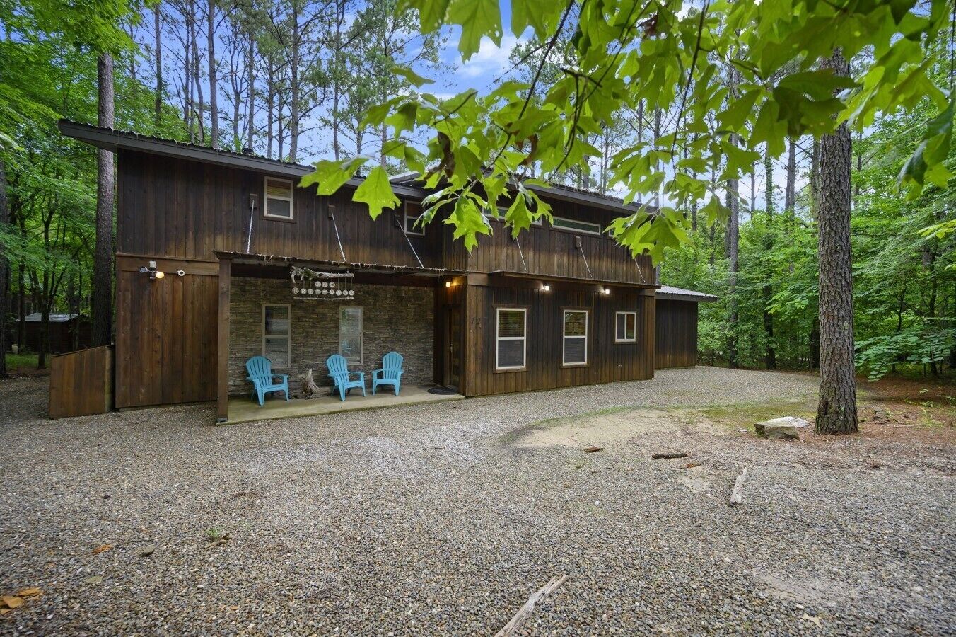 A rustic cabin with a gravel driveway, wooden exterior, and three blue Adirondack chairs outside.