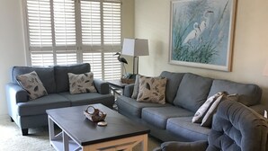 Ein Wohnzimmer mit grauem Sofa, einem passenden Sessel und einem Couchtisch. An der Wand hängt ein Gemälde und auf einem Beistelltisch steht eine Lampe.